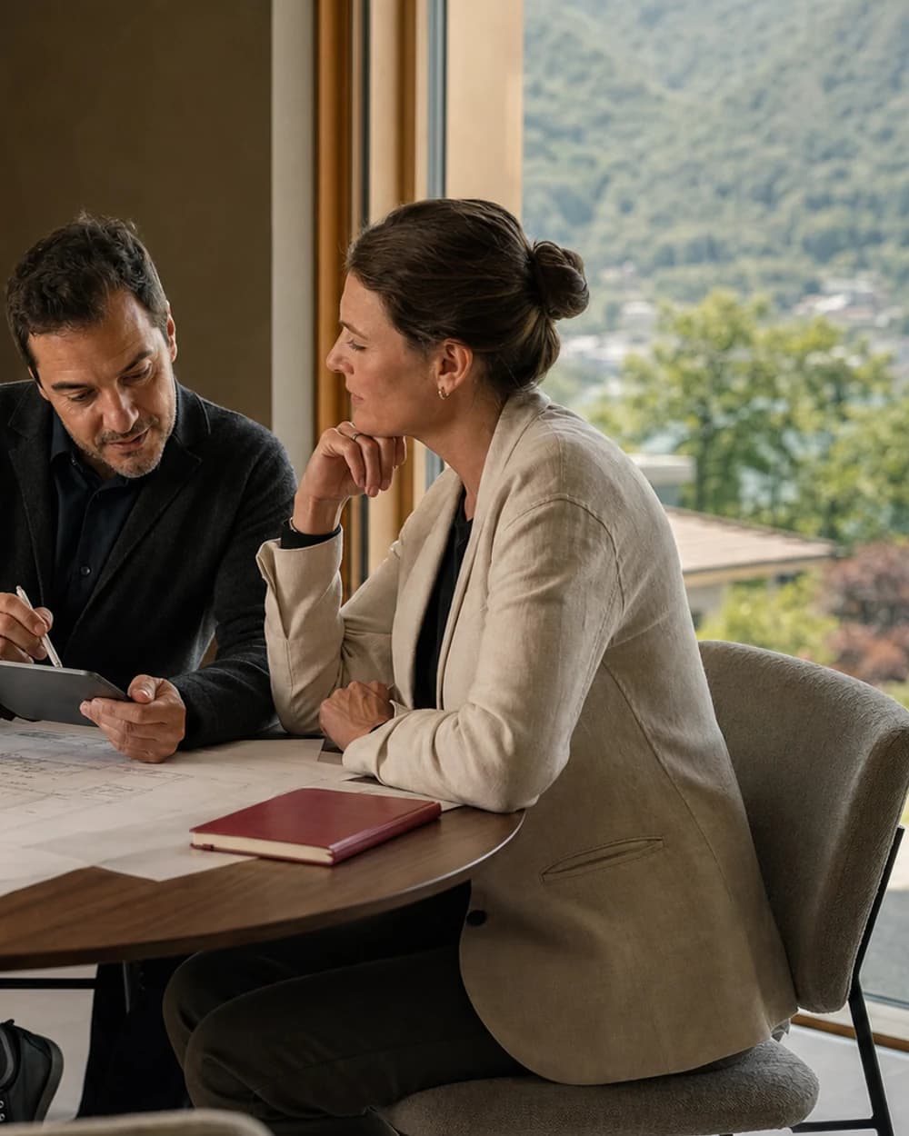 Ein Mann und eine Frau besprechen an einem Tisch Pläne und ein Tablet. Durch das Fenster sind See und bewaldete Berge zu sehen.