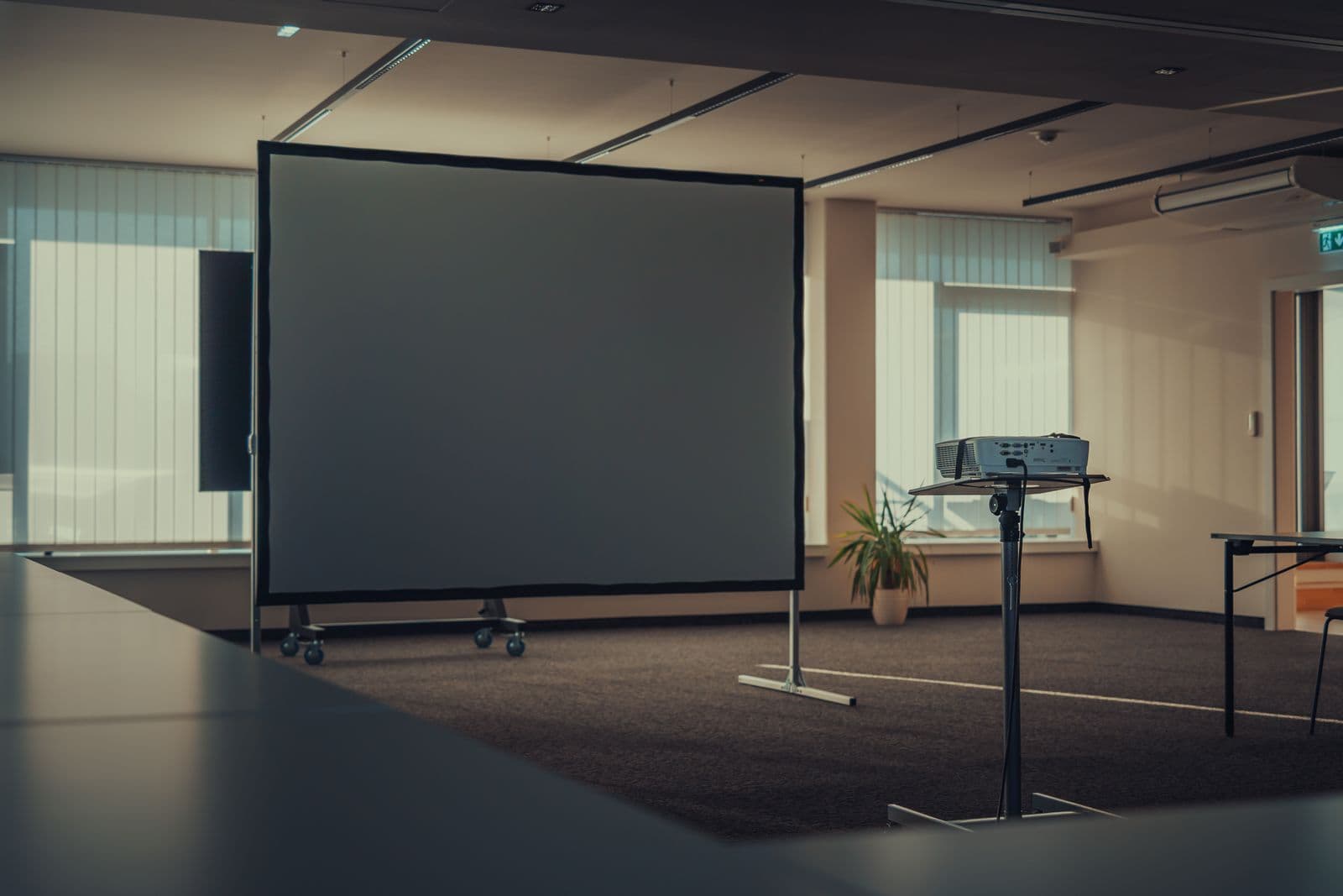 Aufgeräumter Präsentationsraum mit Leinwand und Beamer, beleuchtet vom Tageslicht durch die Lamellenstoren.