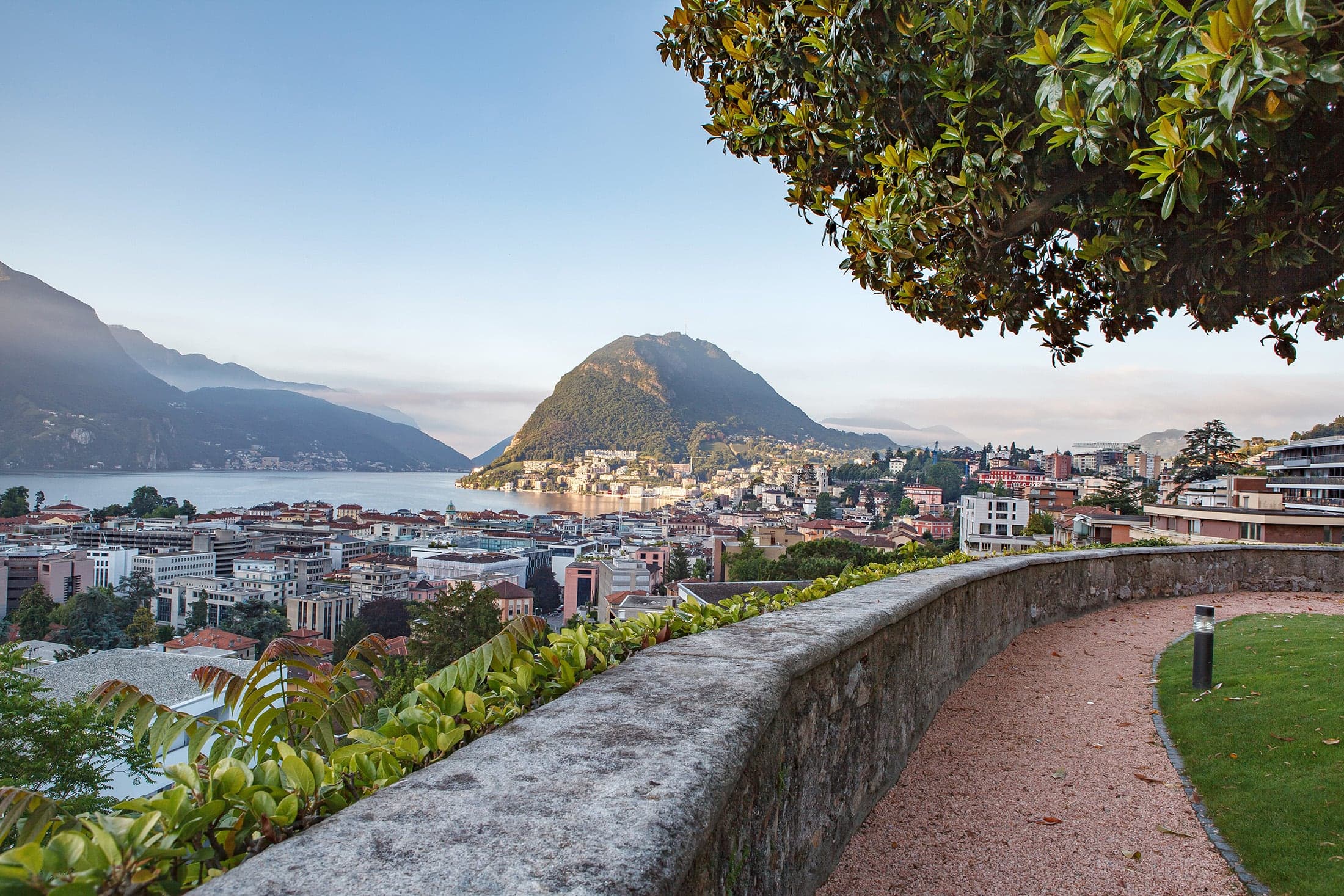Blick über eine Gartenterrasse auf Lugano, den See und den Monte San Salvatore.